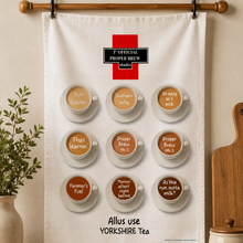 Load image into Gallery viewer, A white fabric tea towel hanging from a wooden rod on a wall, featuring 12 illustrated cups of tea labeled with different tea blends and the text '1st Official Proper Brew Teas' and 'Allus use YORKSHIRE Tea'. The scene is styled in a kitchen-like setting with a plant on the left and a wooden cutting board and jar on the right.