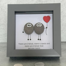 Load image into Gallery viewer, A handmade frame with natural pebbles depicting two cartoon figures holding a red heart balloon, with a white background and the phrase 'There are friends, there is family, and there are friends that become family' printed at the bottom.