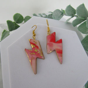 Pink lightning bolt earrings with gold accents on a white card with green leaves in the background