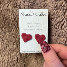Load image into Gallery viewer, Red heart-shaped leather earrings on a Shadow Crafts card against a textured brown background.
