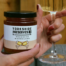 Load image into Gallery viewer, Person holding a jar of Yorkshire Preserves Prosecco Marmalade and a glass of white wine.
