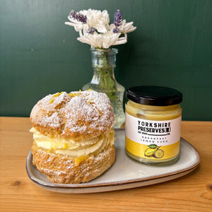 Pastry with lemon curd and cream on a plate next to a jar of Yorkshire Preserves on a wooden surface.