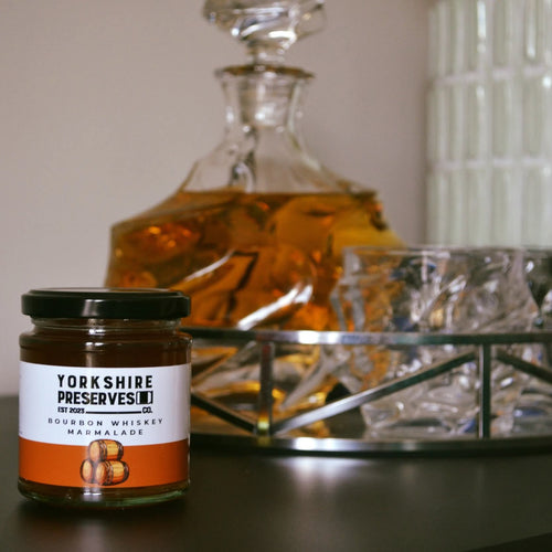 Jar of Yorkshire Preserves' bourbon whiskey marmalade with a decanter and glass in the background.