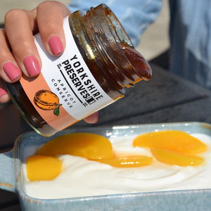 Person pouring apricot preserve from a jar labeled 'Yolkshun Preserves' onto yogurt with peaches.
