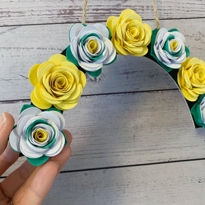 Decorative arch made of paper flowers in yellow, green, and white held by a hand on a wooden surface.