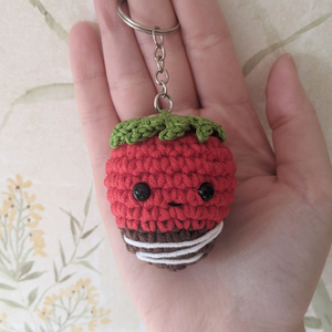 Hand holding a small crochet strawberry keychain with a floral background