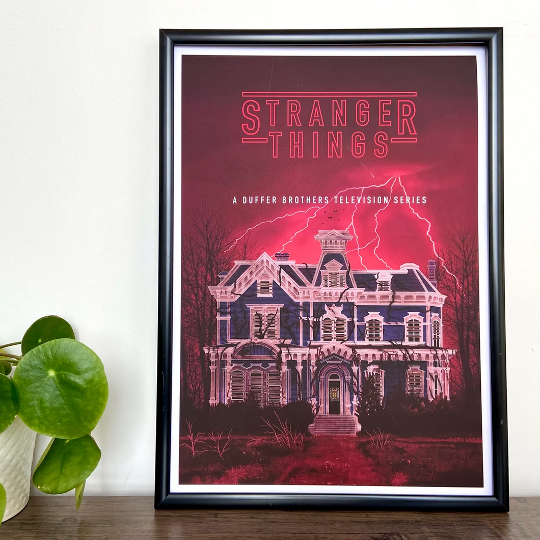 Framed 'Stranger Things' poster with a house illustration on a red background, placed on a wooden surface with a plant.